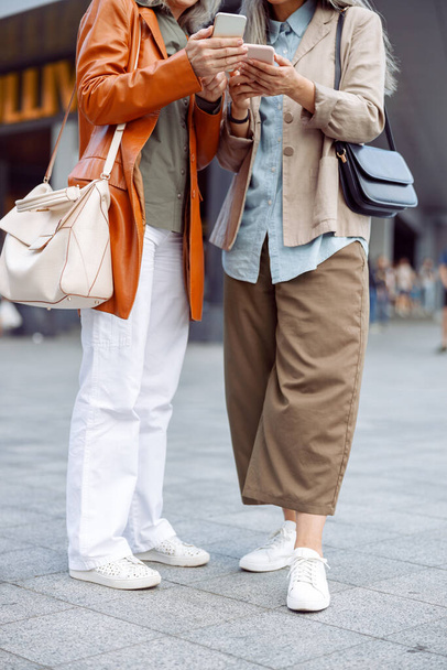 Mature women in stylish apparel hold modern mobile phones standing on city street - Photo, Image