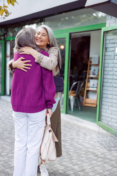 Ευτυχισμένη ηλικιωμένη Ασιάτισσα αγκαλιάζει καλύτερα φορώντας μωβ σακάκι στο δρόμο της πόλης - Φωτογραφία, εικόνα