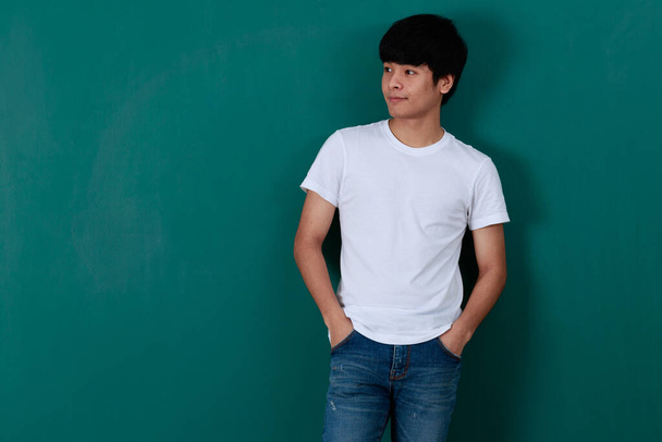 Zachwycony młody azjatycki mężczyzna w białej koszuli i dżinsy stojące na zielonym tle w studio i patrząc puste miejsce z confedent. - Zdjęcie, obraz