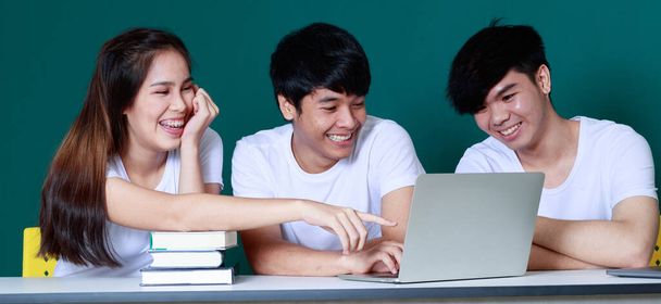 スタジオで緑の背景で一緒に試験の準備をしながら、本やノートパソコンでテーブルに座っているアジアの学生の陽気な会社 - 写真・画像