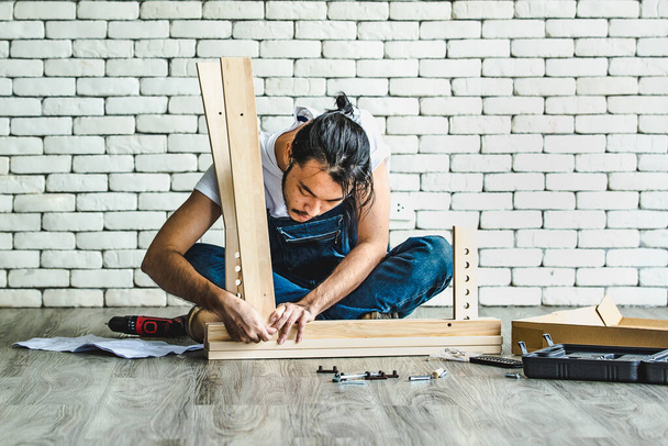 Hipster fiatalember dolgozik ezermester, összeszerelése fa asztal felszereléssel és kézi, koncepció otthoni diy és önkiszolgáló fehér tégla blokk fal a háttérben. - Fotó, kép