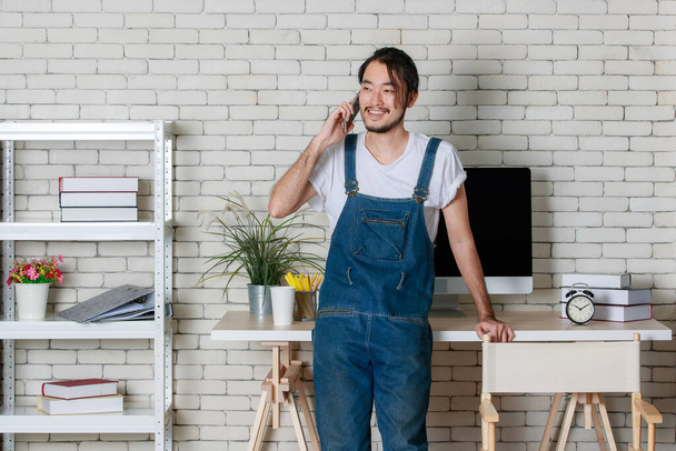 Joven barba hipster hombre asiático de pie puede hacer una llamada en el teléfono inteligente con feliz en su nueva oficina de inicio mínimo moderno. - Foto, Imagen