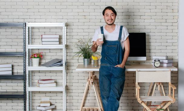 Młody hipsterski broda Azjata stojący ze szczęściem w swoim nowoczesnym minimalnym biurze startowym, koncepcja dla szczęśliwego biznesmena z małym biznesem zaczynającym. - Zdjęcie, obraz