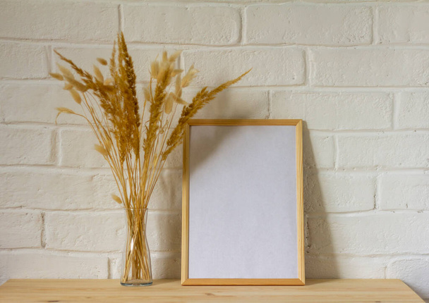 Uma maquete de uma moldura de fotos vazia e um buquê de outono de ervas secas em um vaso transparente em uma mesa de madeira com uma cópia do espaço - Foto, Imagem