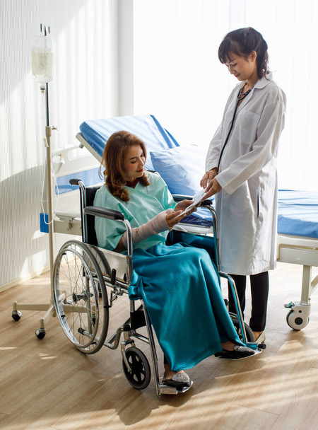 Γυναίκα ασθενής κάθεται σε αναπηρική καρέκλα και να επικεντρωθεί ακρόαση, ενώ ο γιατρός δείχνουν έκθεση ατυχήματος επίσης να εξηγήσει πώς να ανακτήσει το χέρι της σπασμένα από την επαγγελματική υποστήριξη - Φωτογραφία, εικόνα