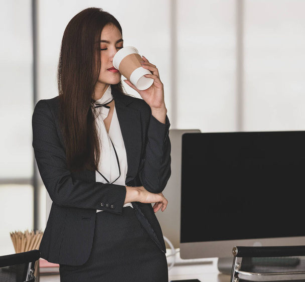 Young attractive Asian woman in black business drinking a cup of coffee in modern looking office with blurry windows background. Concept for modern office lifestyle. - Photo, Image