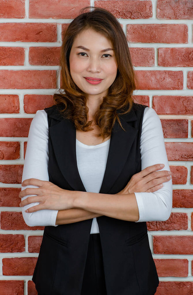 Retrato de meia-idade atraente mulher asiática em colete preto e t-shirt branca posando e sorrindo para a câmera com fundo de parede de tijolo laranja. - Foto, Imagem