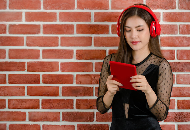 Ritratto di giovane bella attraente asiatica modello femminile in abito nero rivelando indossare cuffie rosse ascoltare musica e guardando tablet contro sfondo muro arancio mattone. - Foto, immagini