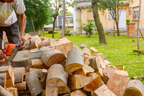 Ο ξυλοκόπος κόβει κορμούς δέντρων στην αυλή χρησιμοποιώντας επαγγελματικό αλυσοπρίονο και τα πετάει στη στοίβα.. - Φωτογραφία, εικόνα
