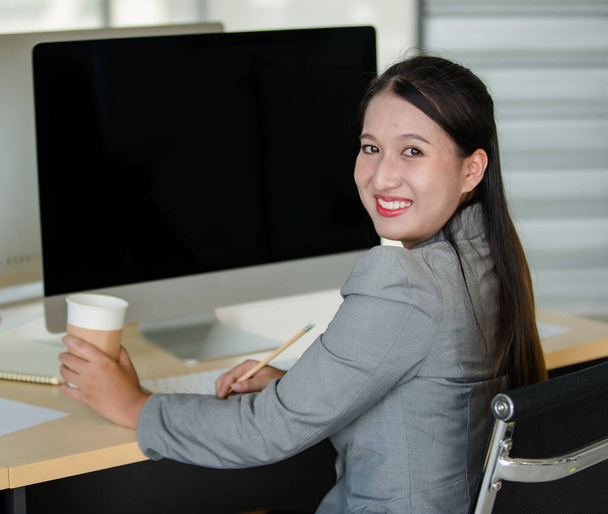 Young attractive Asian woman in grey business suit sitting holding pencil in modern looking office with blurry windows background. Concept for modern office lifestyle. - Foto, Bild