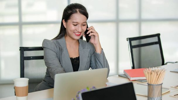 Mladá atraktivní asijská žena v šedém obleku sedí a mluví na mobilním telefonu v moderně vypadající kanceláři s rozmazanými okny pozadí. Koncept pro moderní kancelářský životní styl. - Fotografie, Obrázek