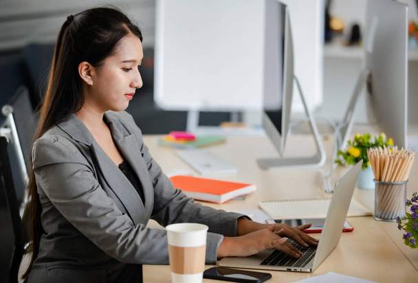 Giovane donna asiatica attraente in abito grigio che lavora sul computer portatile in ufficio dall'aspetto moderno. Concetto per uno stile di vita moderno. - Foto, immagini