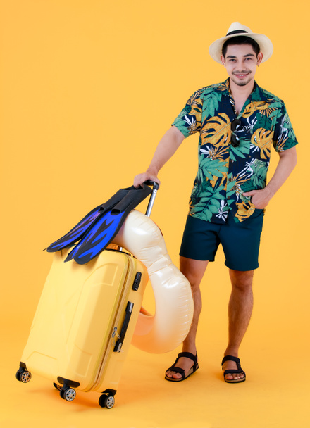Hezký mladý Asiat drží žlutý kufr, ploutve a plavecký prsten. Full body studio portrét na žlutém pozadí. Koncept přípravy letních prázdnin - Fotografie, Obrázek