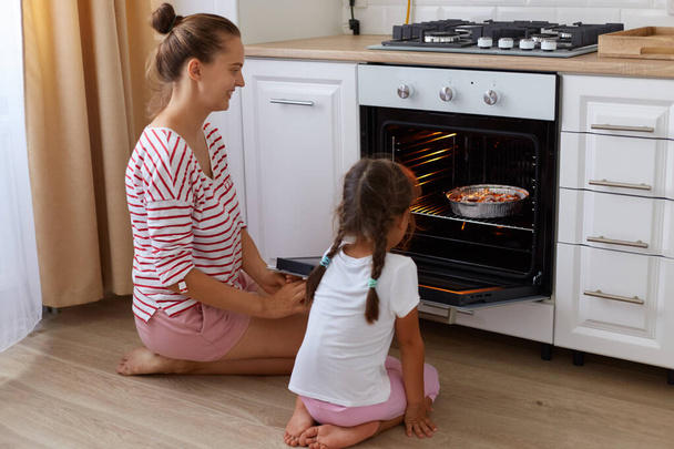 Side view portrait of woman wearing striped casual shirt and her daughter in white t shirt sitting on floor in kitchen and waiting their baking being ready. - Foto, Imagen