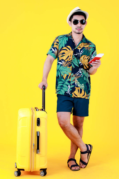 Enjoyed young Asian man in colorful Hawaiian shirt holds and point finger to  passport near suitcase. Full body studio portrait on yellow background. Happy summer vacation travel concept - Foto, Imagen