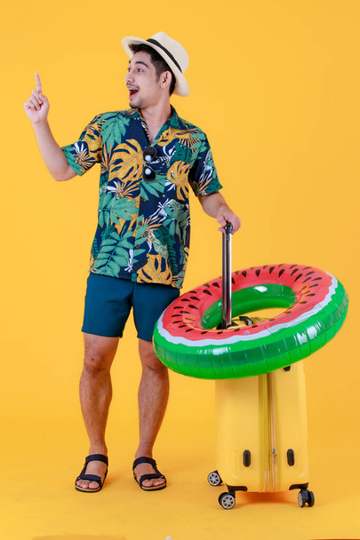 Profilbild, junger asiatischer Mann in buntem Hawaiihemd reicht Zeigefinger nach oben und zieht gelben Koffer. Ganzkörper-Studioporträt auf gelbem Hintergrund. Glückliches Sommerferienreisekonzept - Foto, Bild