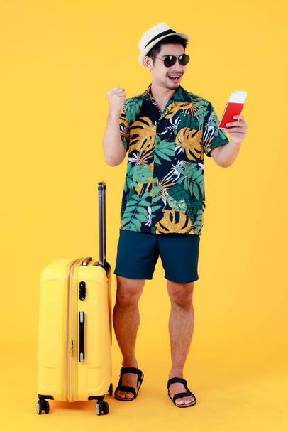 Renkli Hawaii tişörtlü, neşeli Asyalı genç adam pasaport tutuyor ve valizin yanında güzel bir jest yapıyor. Sarı arka planda tüm vücut portresi. İyi tatiller. - Fotoğraf, Görsel