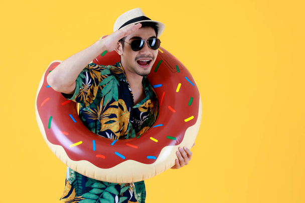 Stylischer junger asiatischer Mann mit Sonnenbrille hält bunten Schwimmring in der Hand. Studioporträt auf gelbem Hintergrund. Sommerferien starten Konzept - Foto, Bild