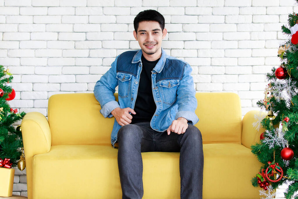 Trendy handsome Asian man sits on yellow sofa near decorated Christmas trees. Portrait of fashionable guy during Christmas and New Year holidays Concept - Photo, Image