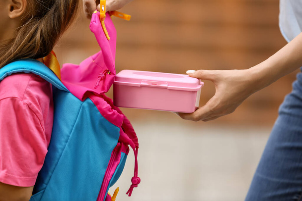 Мама кладет школьный обед в рюкзак своей маленькой дочери на открытом воздухе - Фото, изображение