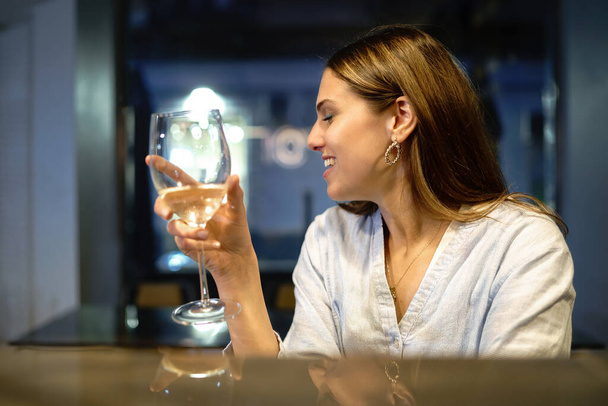 Młoda uśmiechnięta jasnobrązowa kobieta ubrana w bluzkę patrzy na kieliszek białego wina siedzącego przy ladzie baru restauracyjnego. - Zdjęcie, obraz