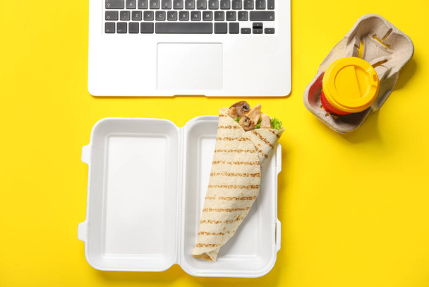 Boîte à lunch en plastique avec Fajitas savoureux, ordinateur portable et tasse de café sur fond jaune - Photo, image