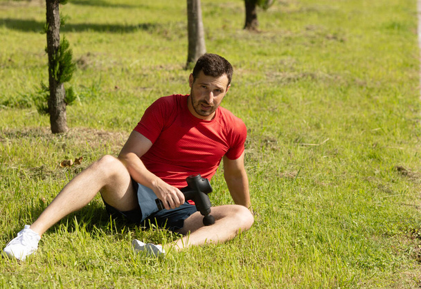 athletic man sitting on grass and massaging body with massage percussion device after workout. - Foto, imagen