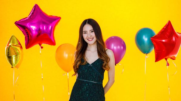 Menina feliz asiática usa vestido sexy em pé posando e divertido animado com papel brilhante confete na mão na festa de celebração na frente amarelo parede fundo e coloridos balões de ar de hélio. - Foto, Imagem