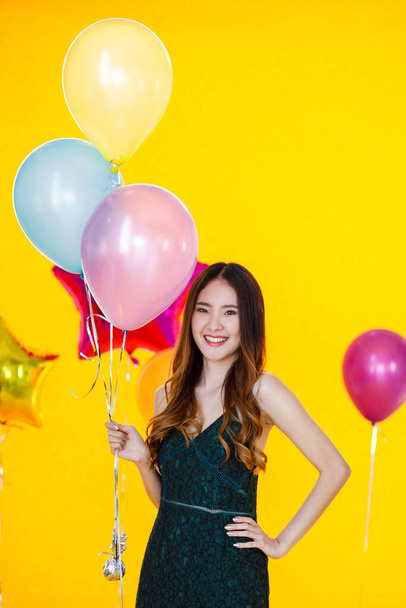 Portrait studio shot of Asian happy beautiful party girl in sleeveless dress stand smiling holding colorful helium air balloons look at camera in front yellow background in new year celebration event. - Valokuva, kuva
