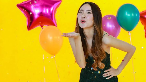 Asian happy girl wears sexy dress standing posing and fun excited with shiny paper confetti in hand in celebration party in front yellow wall background and colorful helium air balloons. - Photo, Image