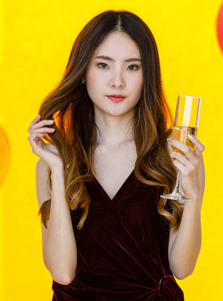 Detailní záběr asijské docela dlouhé vlasy dívka v sexy šaty stojící popíjení červený koktejl nápoj alkohol nápoj ve vysoké sklo v přední barevné balónky na rozmazané žluté pozadí ve straně. - Fotografie, Obrázek