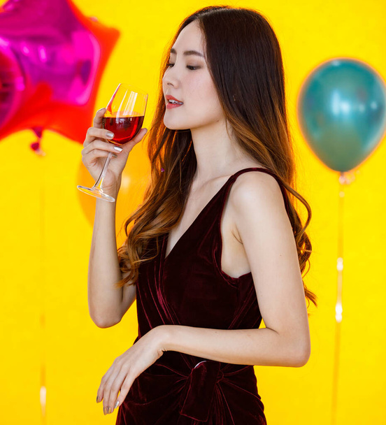 Asian beautiful confident girl in sexy dress standing crossed arm holding cheers toasting red wine beverage alcohol drink in tall glass in front colorful balloons on blurred yellow background party. - Photo, Image