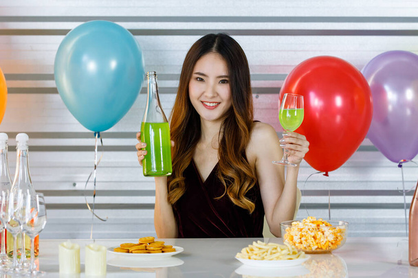 Asiática hermosa chica en vestido sexy sentado sonriendo verter verde manzana martini cóctel de botella en copa de vino alto en fiesta en la mesa de alimentos y bebidas en frente globos de colores en el fondo. - Foto, imagen