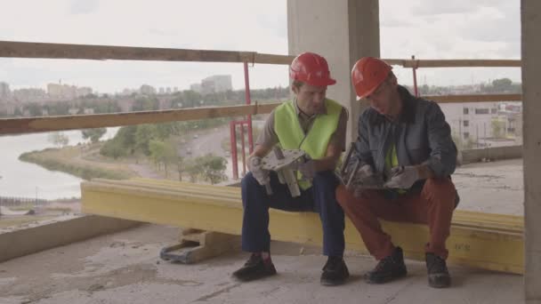 Weitwinkelaufnahme von zwei kaukasischen Männern mit Helmen und Arbeitskleidung, die auf einem Balken im Vordergrund der Stadtansicht sitzen, reden und metallische mechanische Teile in der Hand halten, lächelnd - Filmmaterial, Video