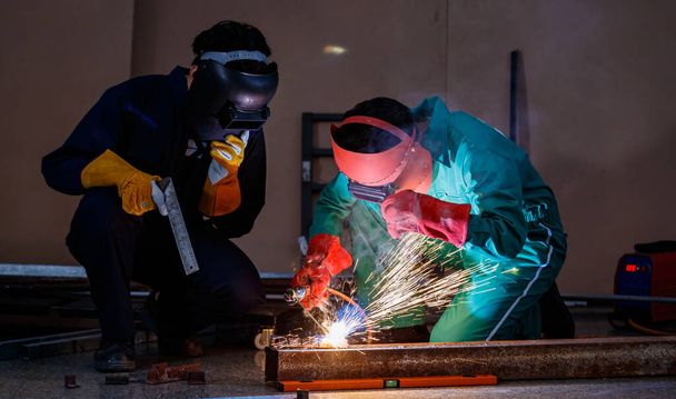 Two engineers mechanics sitting and working in a workshop of a factory. They are helping each other to weld a piece of metal rod with a welding machine at night time. - Photo, Image