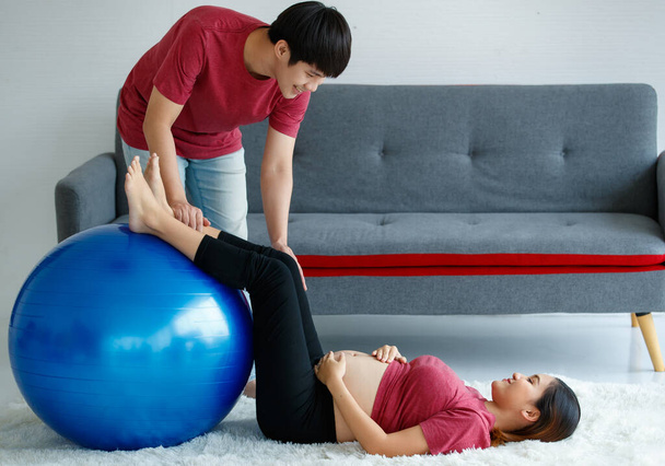 Joyeux beau jeune homme asiatique assis regardant une femme enceinte joyeusement. mari actif encourage sa femme à faire de l'exercice avec un ballon de pilates à la maison. - Photo, image