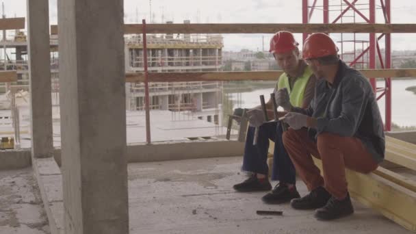 Lange Aufnahme von zwei kaukasischen Arbeitern mit Helmen, die auf einem Balken im Vordergrund der Stadtansicht sitzen, reden und metallische mechanische Teile in der Hand halten, lächelnd - Filmmaterial, Video