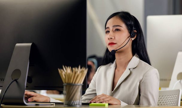Сосредоточьтесь на взрослой азиатской женщины справочной службы поддержки намерены с ее работой носить наушники и смотреть на компьютер монитор карандаш в коробке на рабочем столе. - Фото, изображение