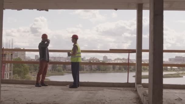 Широкий след слева от двух кавказских рабочих в шлемах, стоящих на краю строящегося здания, разговаривающих в летний день - Кадры, видео