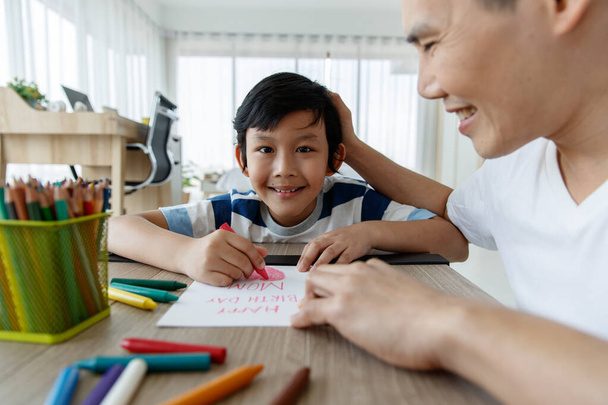 Asiática familia padre e hija está enseñando a mis hijos a escribir tarjetas de felicitación para mamá con hijo pequeño en silla en casa en el día de la madre - Foto, imagen