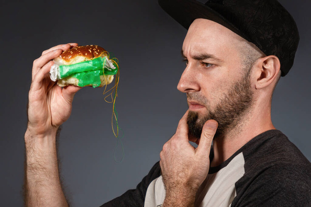 The concept of fast food and ecology. A man in a cap and a beard, holding a hamburger, and cautiously examines its plastic filling. Black background. Close up. - Photo, Image