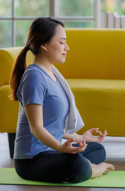 Spor takım elbiseli genç Asyalı kadın yoga yaptıktan sonra gözlerini kapatıp meditasyon yaparken omzuna beyaz havlu koydu. Eğlence ve rahatlama konsepti. - Fotoğraf, Görsel