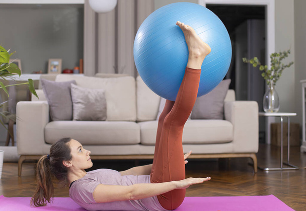 Young woman exercising at home in a living room. She is exercising with a fitness bal - Фото, изображение