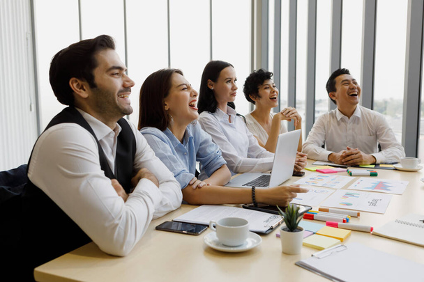 Группа из пяти бизнесменов, двух мужчин и трех женщин, сидящих за столом заседаний и обратите внимание с довольным смехом на спикера конференции - Фото, изображение