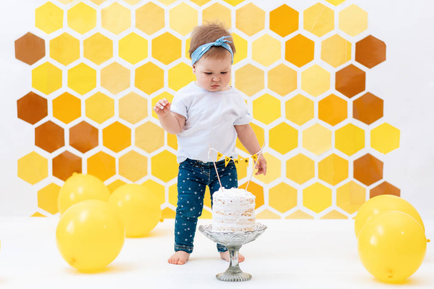 Ein süßes kleines Mädchen steht neben einer Torte. Im Hintergrund ein Design aus gelben Waben und Luftballons. Smash-Kuchen-Konzept. - Foto, Bild