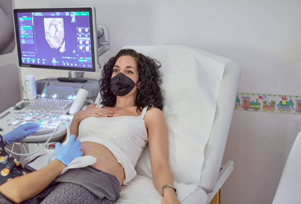 婦人科超音波検査を受けているコロナウイルスパンデミックのため顔マスクをした若い妊婦。妊娠の概念的妊娠のケアと制御 - 写真・画像