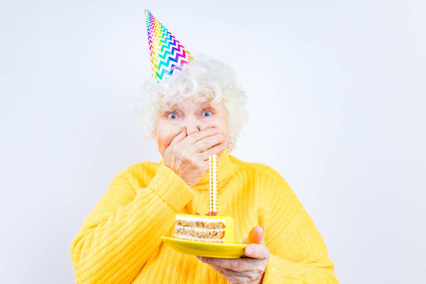 older woman with a gift wear yellow sweater and horn cap on a white background holding plate with cake with fireworks - Foto, Imagem