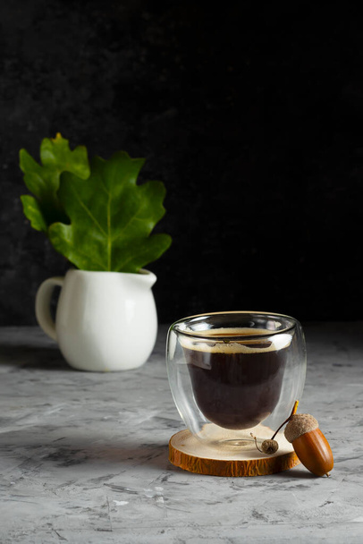 Double bottom glass cup with coffee on wooden stand with acorn and white milk jug with green oak leaves in low key - Φωτογραφία, εικόνα