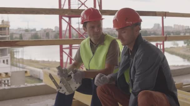 Mittlere Einstellung von zwei kaukasischen Bauarbeitern mit orangefarbenen Helmen, die auf einem Balken im Gebäude im Vordergrund der Stadtansicht sitzen, reden und metallische mechanische Teile in der Hand halten, lächelnd - Filmmaterial, Video