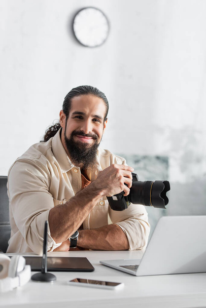 szczęśliwy fotograf z aparatem cyfrowym siedzi w pobliżu zamazanego laptopa i tabletu graficznego w domu - Zdjęcie, obraz